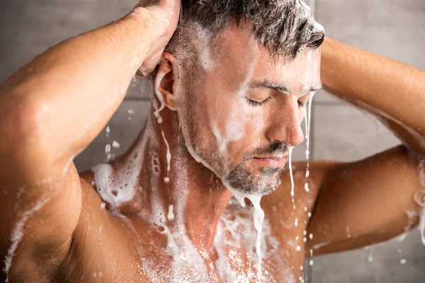 Selective focus of adult man with closed eyes washing foam in shower — Stock Photo
