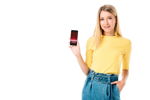 Hermosa mujer joven sosteniendo teléfono inteligente con cursos de comercio en la pantalla y sonriendo a la cámara aislada en blanco - foto de stock