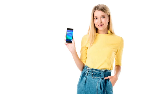 Hermosa mujer joven sosteniendo teléfono inteligente con aplicación de reserva en la pantalla y sonriendo a la cámara aislada en blanco - foto de stock