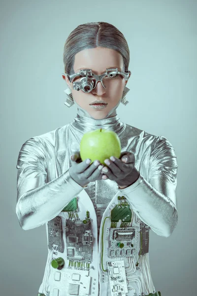 Киборг, держащий зеленое яблоко и смотрящий на камеру, изолированную на сером, концепция технологии будущего — стоковое фото
