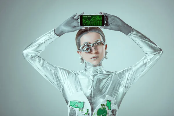 Robot celebración de teléfono inteligente con aplicación de comercio en línea y mirando a la cámara aislada en gris, concepto de tecnología futura - foto de stock