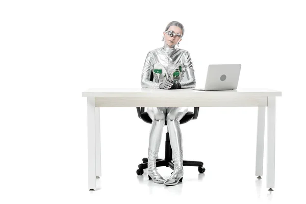 Robot argento seduto a tavola con computer portatile isolato su bianco, concetto di tecnologia futura — Foto stock