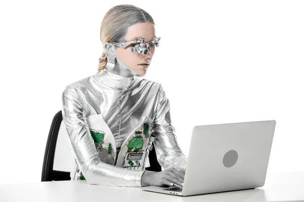 Silver robot sitting at table and working with laptop isolated on white, future technology concept — стоковое фото