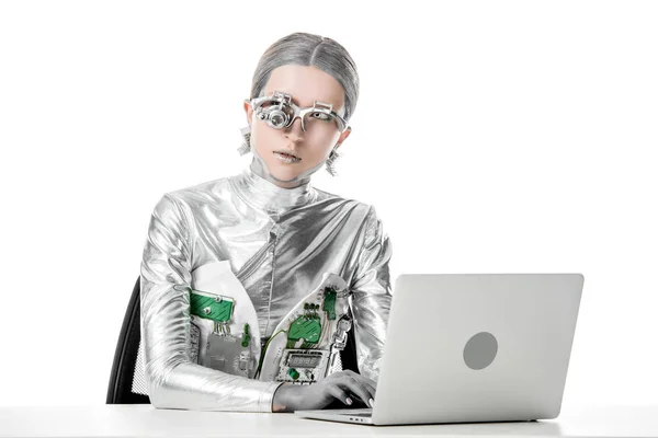 Robot d'argento seduto a tavola con computer portatile e guardando altrove isolato sul bianco, concetto di tecnologia futura — Foto stock