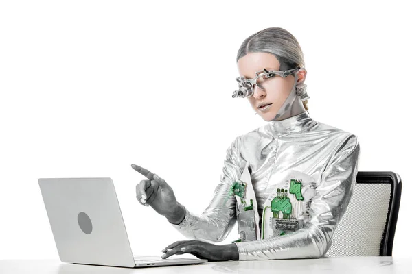 Silver robot sitting at table with laptop and pointing on something isolated on white, future technology concept — стоковое фото
