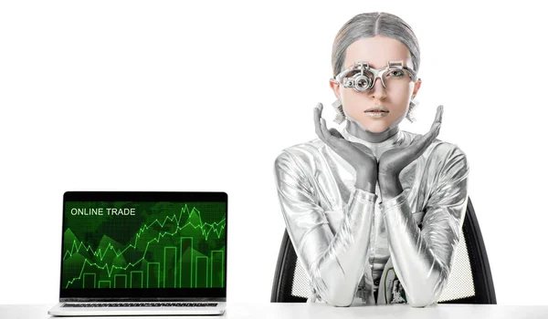 Silver robot sitting at table near laptop with online trade appliance isolated on white, future technology concept — Stock Photo