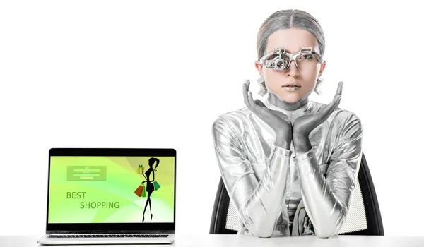 Robot plateado sentado en la mesa cerca de la computadora portátil con el mejor aparato de compras aislado en el concepto de tecnología blanca, futuro - foto de stock