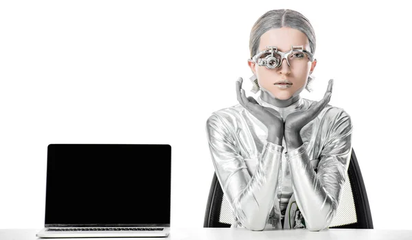 Silver robot sitting at table near laptop with blank screen isolated on white, future technology concept — Stock Photo