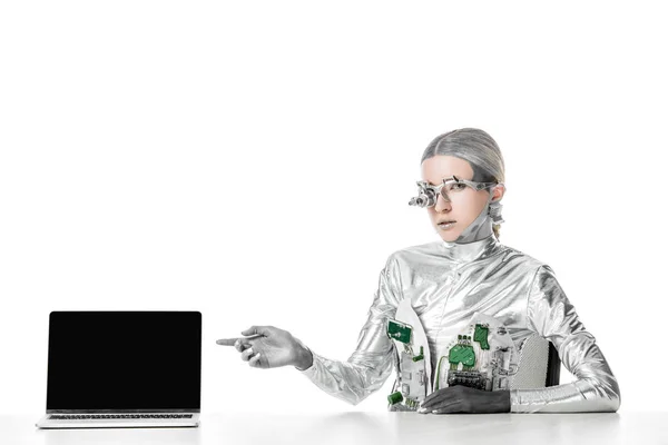 Robô de prata sentado à mesa e apontando no laptop com tela em branco isolado em branco, conceito de tecnologia futuro — Fotografia de Stock