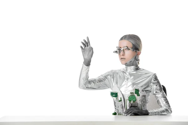 Silver robot with eye prosthesis sitting at table and touching something isolated on white, future technology concept — Stock Photo