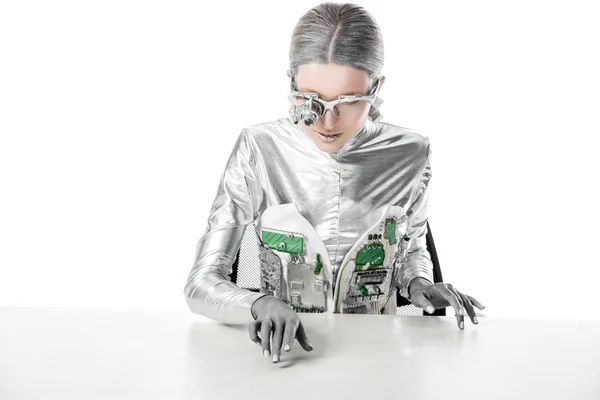 Silver robot with eye prosthesis sitting at table and touching something on tabletop isolated on white, future technology concept — Stock Photo