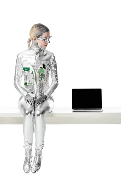 Robot argenté assis sur la table près d'un ordinateur portable avec écran blanc isolé sur blanc, concept technologique futur — Photo de stock
