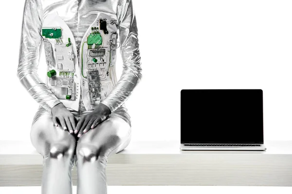 Обрезанное изображение робота, сидящего на столе рядом с ноутбуком с чистым экраном, изолированным на белом, концепция технологии будущего — стоковое фото