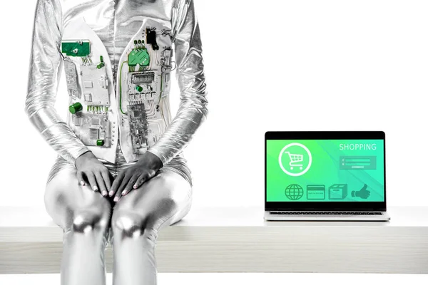 Обрезанное изображение робота, сидящего на столе рядом с ноутбуком с торговым устройством, изолированным на белом, концепция технологии будущего — стоковое фото