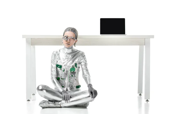 Robot argento seduto vicino al tavolo con computer portatile isolato su bianco, concetto di tecnologia futura — Foto stock