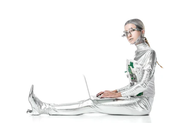 Side view of silver robot sitting with laptop and looking at camera isolated on white, future technology concept — Stock Photo