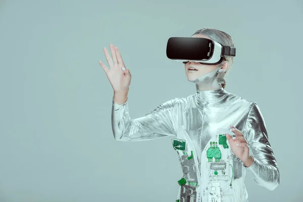 Удивленный серебряный робот, трогающий что-то с помощью гарнитуры виртуальной реальности, изолированной на серой концепции технологии будущего — стоковое фото