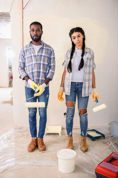 Jeune couple avec brosses à roulettes regardant la caméra pendant la rénovation de la maison — Photo de stock