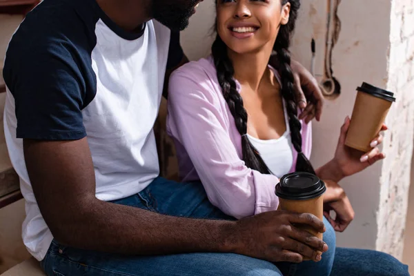 Primer plano de la sonriente pareja sentada en las escaleras con tazas de papel de café durante la renovación del hogar - foto de stock