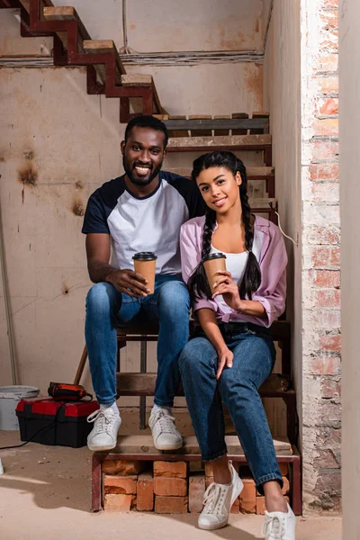 Lächelndes Paar sitzt mit Pappbechern Kaffee auf der Treppe während der Renovierung des Hauses und blickt in die Kamera — Stockfoto