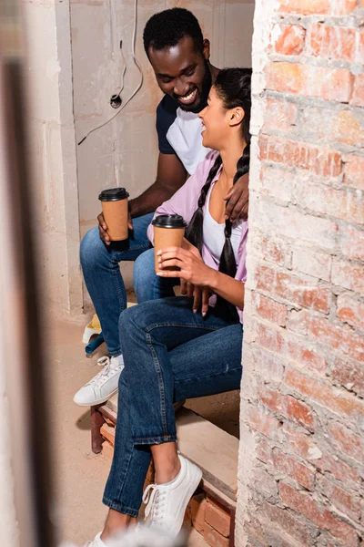 Смеющаяся пара, сидящая на лестнице с бумажными чашками кофе во время ремонта дома — стоковое фото