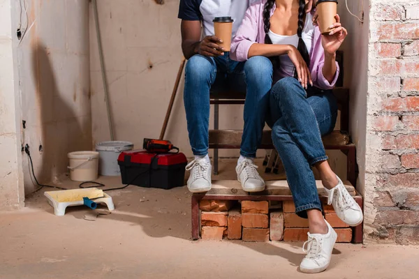 Recortado disparo de pareja sentado en las escaleras con tazas de papel de café durante la renovación - foto de stock