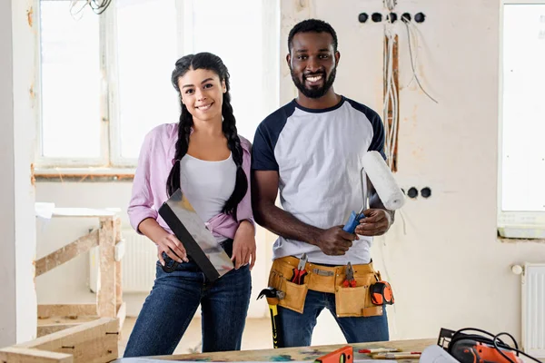 Feliz jovem casal com ferramentas olhando para a câmera ao fazer a renovação de casa — Fotografia de Stock