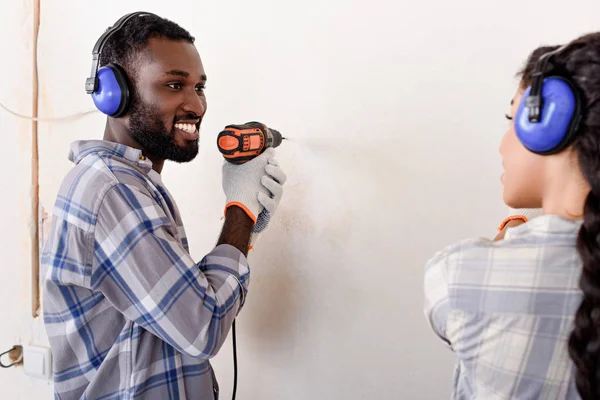 Glücklicher junger Mann, der Wand bohrt und Freundin anschaut, während er Renovierung seines Hauses macht — Stockfoto