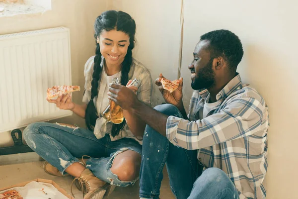 Feliz casal jovem clinking garrafas de cerveja e comer pizza enquanto faz renovação de casa — Fotografia de Stock