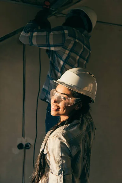 Felice giovane donna in cappello duro guardando la fotocamera mentre suo marito fa la ristrutturazione della casa — Foto stock