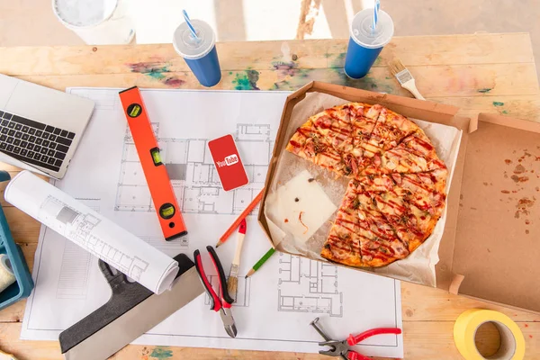 Top view of box with pizza, tools and smartphone with youtube app on screen on building plan — Stock Photo