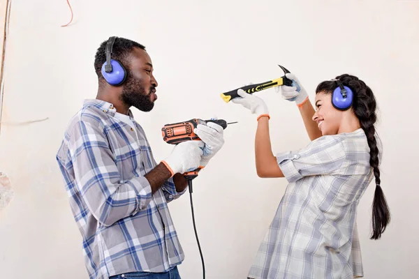 Vista lateral do jovem casal brincalhão se divertindo com martelo e broca ao fazer a renovação de casa — Fotografia de Stock