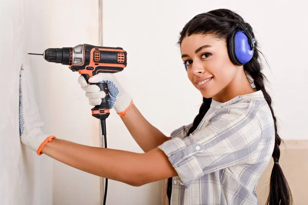 Belle jeune femme forage mur et regarder la caméra pendant la rénovation de la maison — Photo de stock