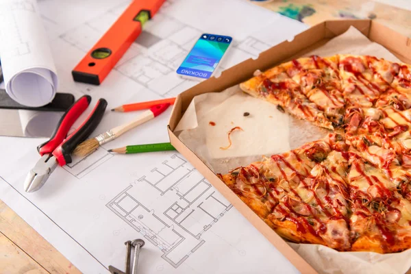 Close-up shot of box with pizza, tools and smartphone with booking app on screen on building plan — Stock Photo