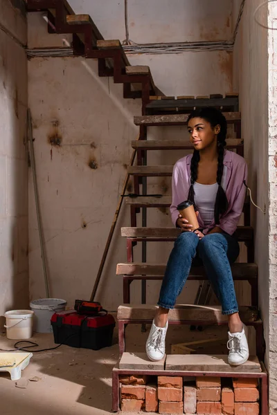 Schöne junge Frau sitzt auf der Treppe mit Pappbecher Kaffee während der Renovierung ihres Hauses — Stockfoto
