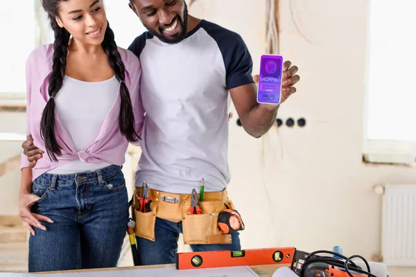 Улыбающаяся пара, держащая смартфон с торговым приложением на экране во время ремонта — стоковое фото