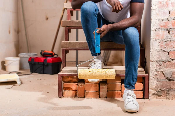 Обрезанный снимок африканского американца, сидящего на лестнице с кисточкой во время ремонта дома — стоковое фото