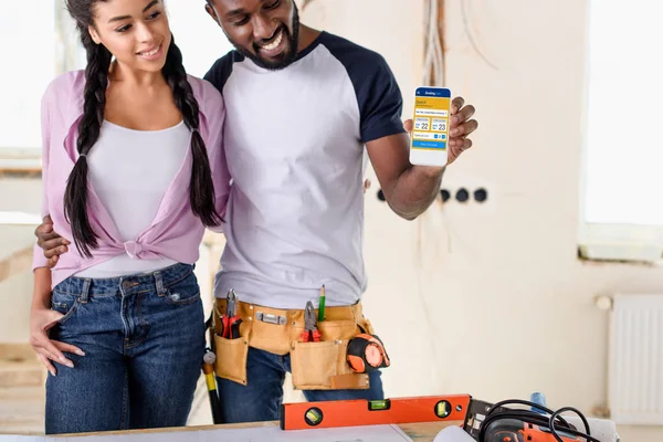 Pareja que sostiene el teléfono inteligente con el sitio web de reserva en pantalla durante la renovación - foto de stock