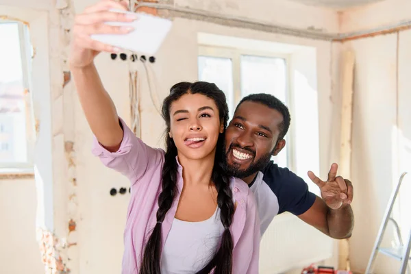 Positivo afroamericano guiño mientras toma selfie con novio haciendo signo de paz en el teléfono inteligente - foto de stock