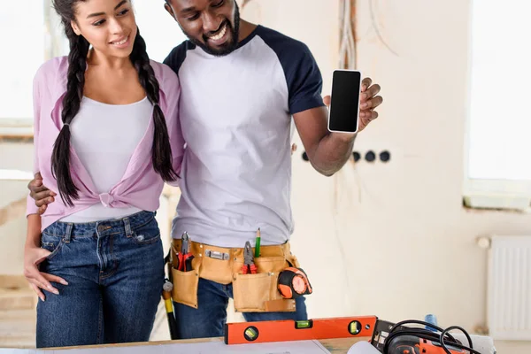 Giovane coppia afro-americana che mostra smartphone con schermo bianco durante la ristrutturazione nella nuova casa — Foto stock