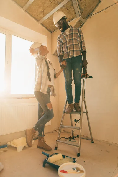 Low angle view of man in hard hat and goggles standing power drill on ladder while his girlfriend standing near during renovation of home — Stock Photo