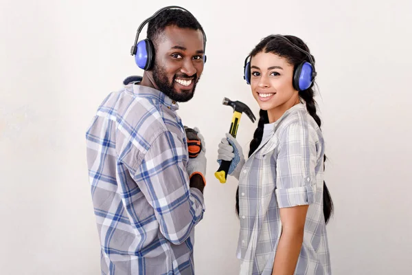 Sonriente pareja afroamericana joven en auriculares protectores de pie con taladro y martillo durante la renovación de un nuevo hogar - foto de stock