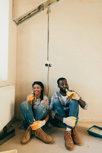 Riéndose pareja afroamericana bebiendo cerveza durante la renovación en casa - foto de stock