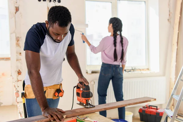 Handsome african american man working with jigsaw while his girlfriend working behind during renovation at home — Stock Photo
