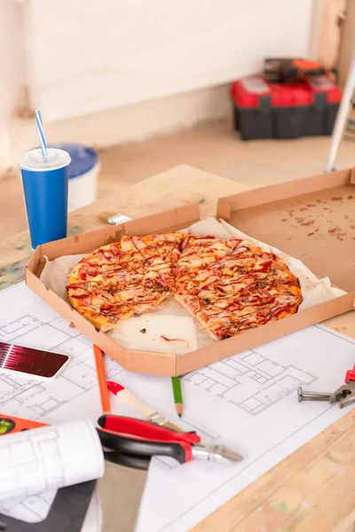 Foco seletivo de pizza, refrigerante, blueprint, ferramentas e smartphone com gráficos na tela na mesa — Fotografia de Stock