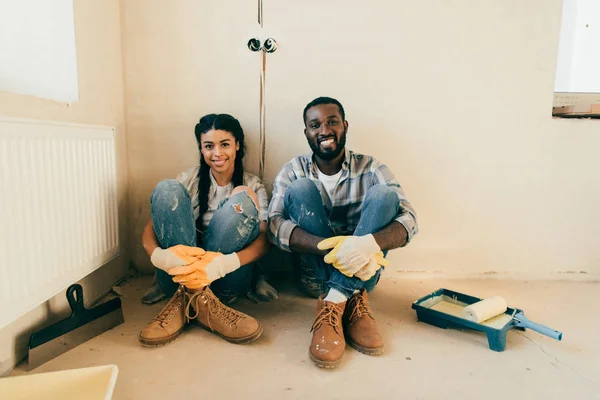 Joyeux couple afro-américain en gants de protection regardant la caméra pendant la rénovation de la maison — Photo de stock