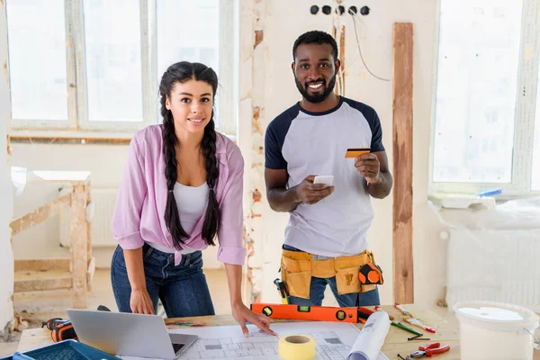 Young african american man with credit card and smartphone standing near girlfriend during renovation of home — Stock Photo