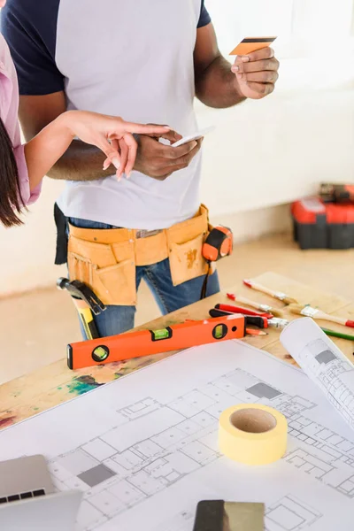 Teilbild einer Frau, die mit dem Finger auf ihr Smartphone zeigt, während ihr Freund bei Renovierungsarbeiten mit Kreditkarte am Tisch mit Blaupause steht — Stockfoto