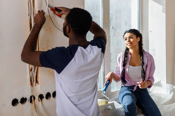 Uomo afro-americano che lavora con le pinze mentre la sua ragazza mette rullo di vernice nel vassoio del rullo sul davanzale della finestra a nuova casa — Foto stock