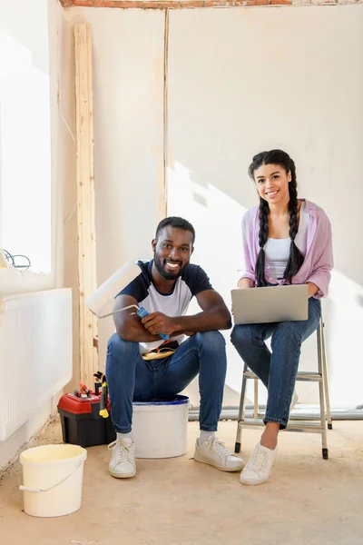 Lächelndes afrikanisch-amerikanisches Paar mit Laptop und Farbroller, das bei Renovierungsarbeiten in die Kamera schaut — Stockfoto